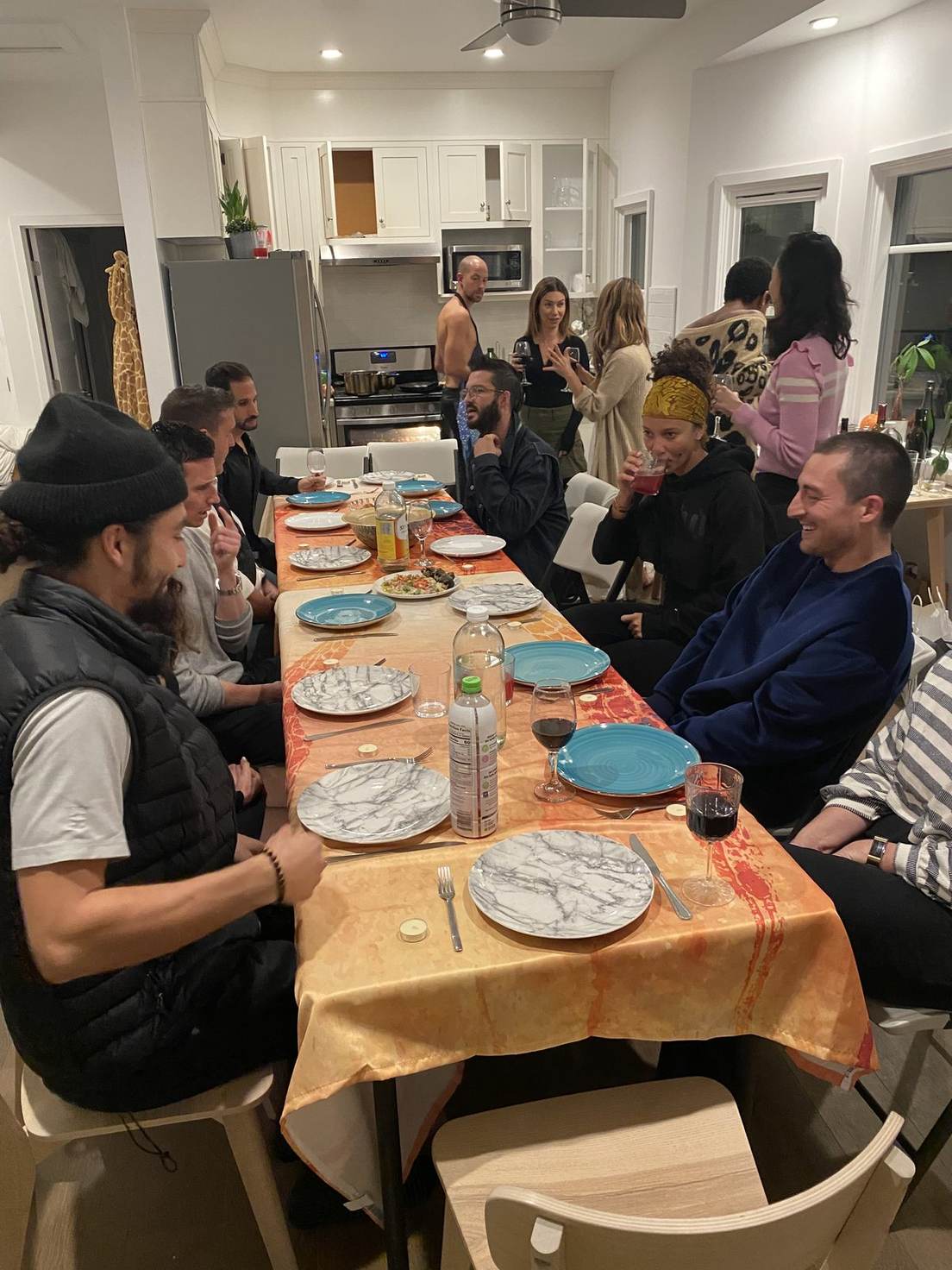 A long dinner table under an orange cloth in a home kitchen, set with marble and turquoise plates and red wine, about fifteen guests seated along both sides and more talking by the stove