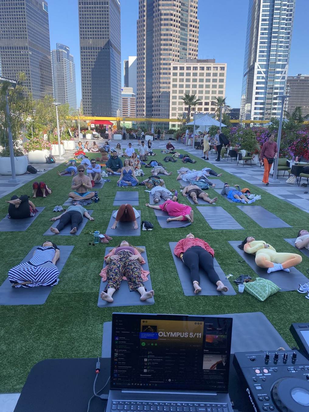 Rows of guests lying on mats on a rooftop lawn between downtown Los Angeles towers at Olympus